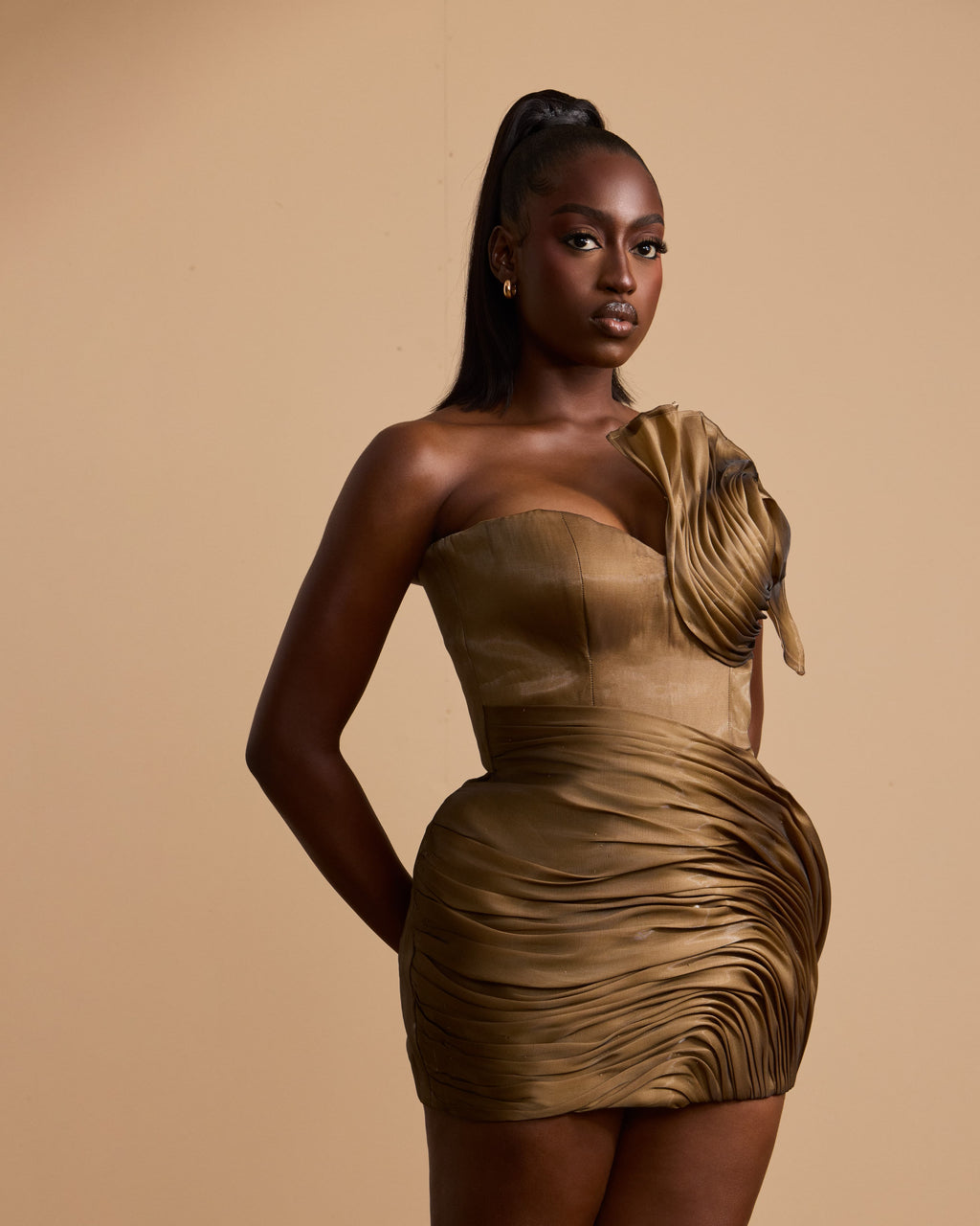 A woman modeling a one-shoulder ruched mini dress in a metallic bronze-gold color, posed against a solid beige studio background.