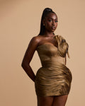 A woman modeling a one-shoulder ruched mini dress in a metallic bronze-gold color, posed against a solid beige studio background.