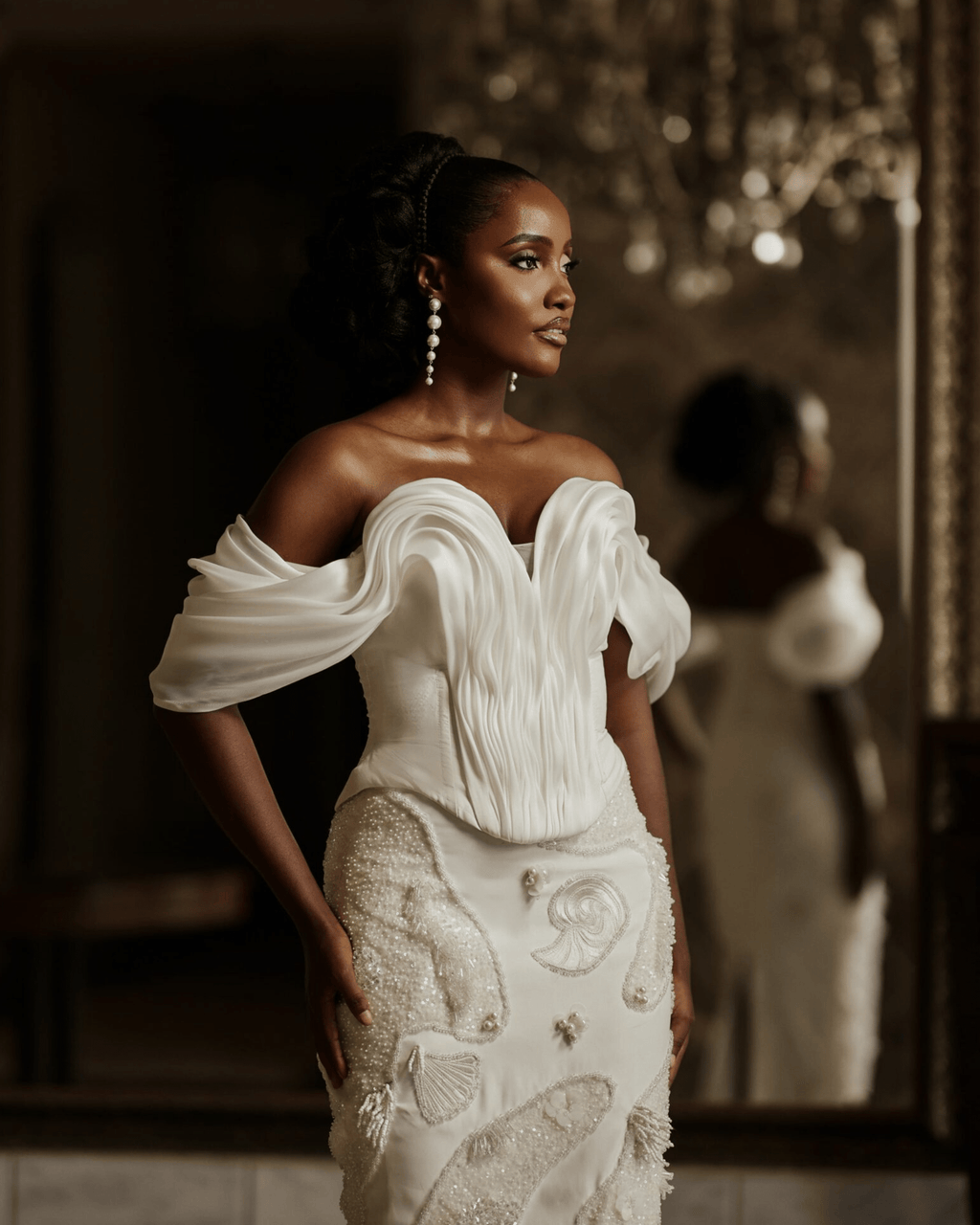 A woman wearing the elegant off-the-shoulder ivory reef top with pleated detailing standing in a dimly lit interior with a mirror reflecting her back.