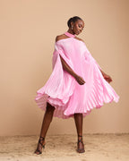 A woman modeling a light pink pleated off-the-shoulder dress with a high-low tiered hem, standing on a sandy surface against a solid beige background.