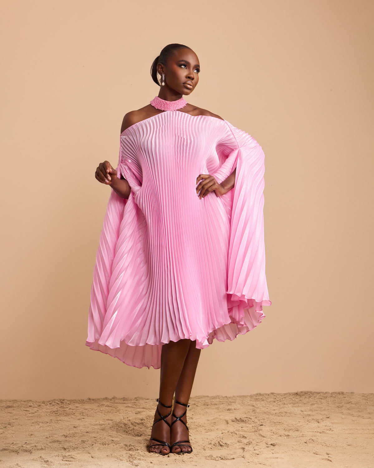 A model wearing a vibrant pink pleated off-the-shoulder dress with a high-low hemline, styled with black strappy heels against a solid beige backdrop.
