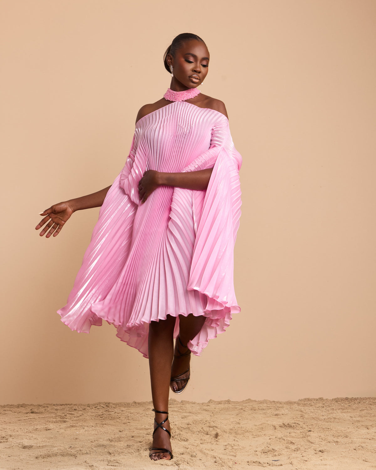 A model wearing a vibrant pink pleated dress with a high-low hemline and draped sleeves, set against a solid beige backdrop.