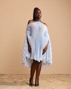 A model wearing a light blue pleated off-the-shoulder cape dress with a high-low hem, styled with a blue beaded choker and black strappy heels, set against a solid beige backdrop.