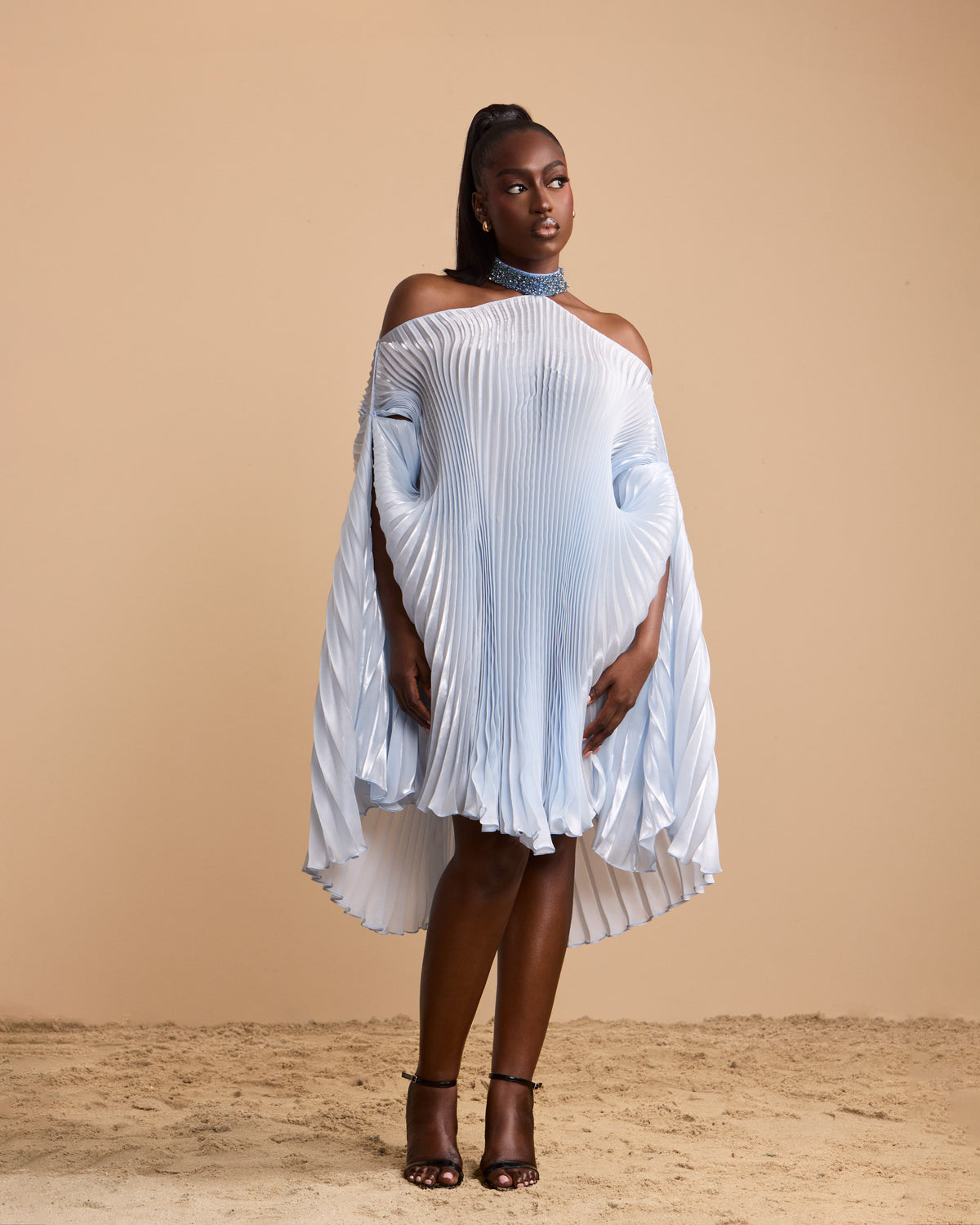 A model wearing a light blue pleated off-the-shoulder cape dress with a high-low hem, styled with a blue beaded choker and black strappy heels, set against a solid beige backdrop.