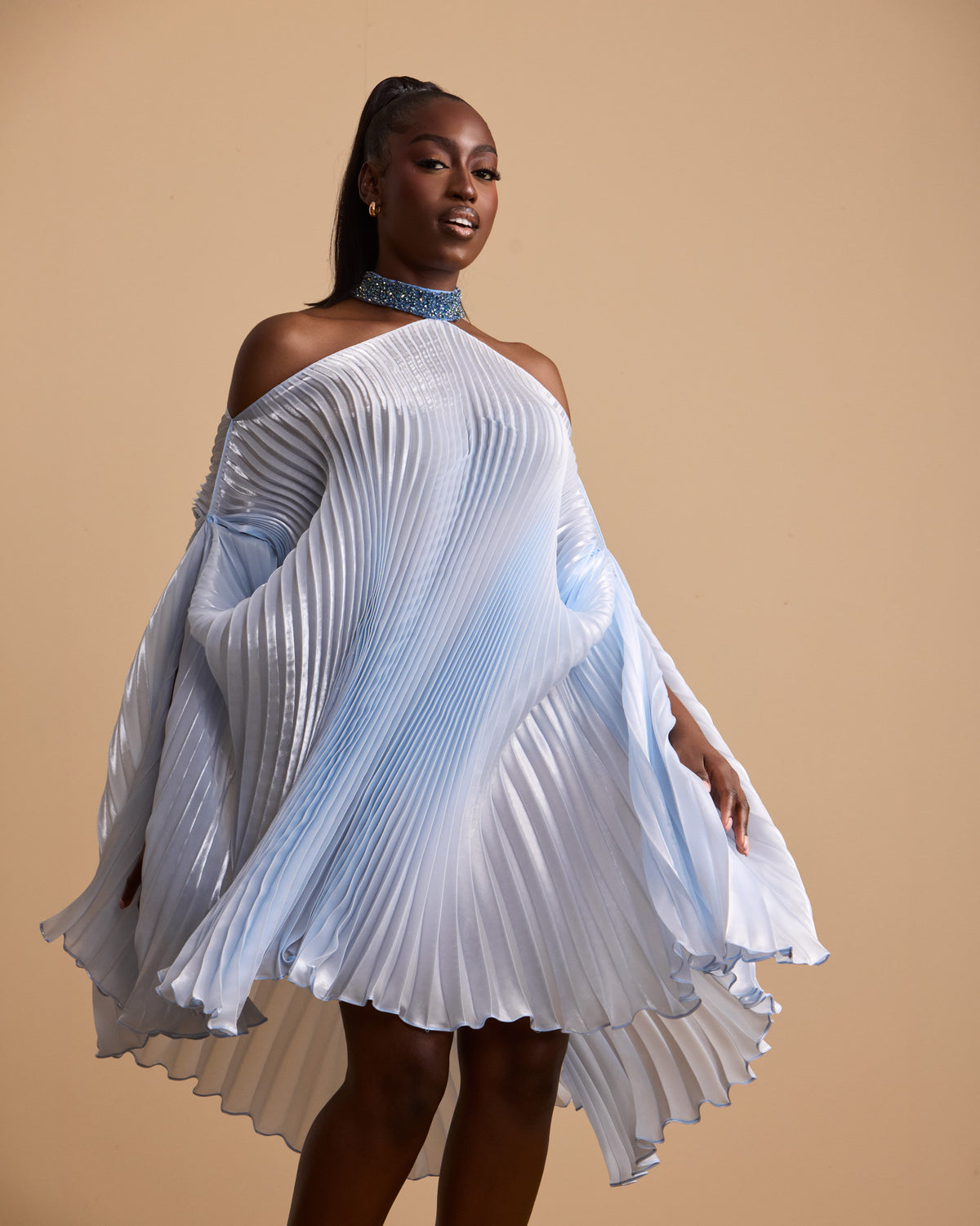 A model wearing a light blue and white pleated halter dress with dramatic draped sleeves, posed against a solid beige studio background.